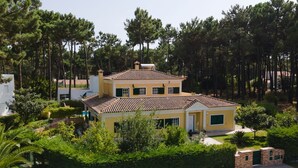 Une maison à deux étages, avec un toit recouvert de tuiles, entourée d’une végétation luxuriante et de grands arbres.