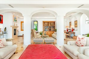 A spacious living room with a red ottoman, white sofas, and a bookshelf.