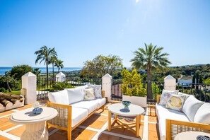 A terrace with white furniture, a view of the sea, and palm trees.
