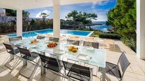 Un espace repas avec une table et des chaises en verre, offrant une vue sur la piscine et l’océan.