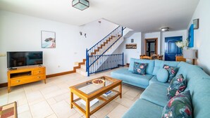 Un salon avec un canapé bleu, une table basse en bois et un téléviseur posé sur un meuble en bois. Un escalier avec des rampes bleues mène à une autre pièce.