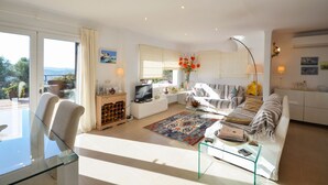 A modern living room with a glass table, a sofa, a TV, and a kitchen area with cabinets and a dining table. - Villa Petunies - Three Bedroom Villa, Sleeps 6 (Begur)