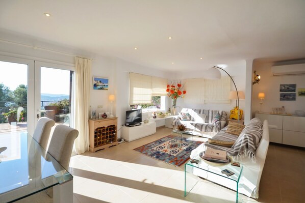 A modern living room with a glass table, a sofa, a TV, and a kitchen area with cabinets and a dining table.