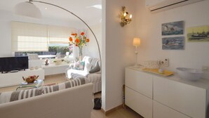 A modern living room with a sofa, a TV, a dining table with a lamp, and a kitchen area with a sink and a bowl. - Villa Petunies - Three Bedroom Villa, Sleeps 6 (Begur)
