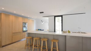 A modern kitchen with wooden cabinets, a large island with stools, a built-in oven, and a window providing natural light. - Villa Falguerines - Six Bedroom Villa, Sleeps 12 (Begur)