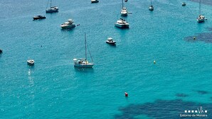 Un puerto deportivo costero con varias embarcaciones fondeadas en aguas tranquilas, rodeado de un paisaje montañoso y un pueblo pequeño al fondo.