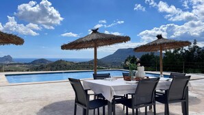 Un área de comedor junto a la piscina con una mesa para dos, dos sombrillas de paja y vistas al mar y a las montañas.