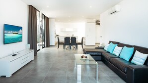 A modern living room with a glass coffee table, a leather sofa with blue cushions, a dining area with a table and chairs, and a kitchen in the background. - Villa Bernialiss - Three Bedroom Villa, Sleeps 6 (Puerto del Carmen)
