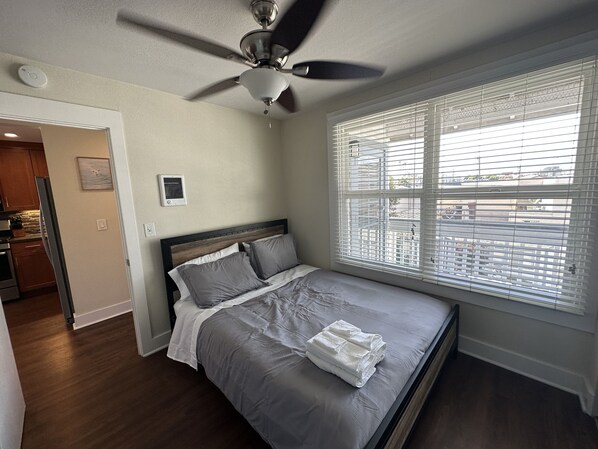 Une chambre avec un lit, un ventilateur de plafond, une fenêtre avec des stores et un coin cuisine visible à travers une porte ouverte.