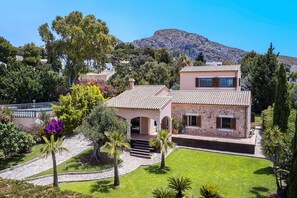 A two-story house with a stone facade, a tiled roof, and a front garden with palm trees and flowering plants.
