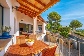A wooden deck with a table and chairs, a bowl of fruit, and a view of a pool and greenery. - Bendinat Golf - Four Bedroom Villa, Sleeps 8 (Calvià)