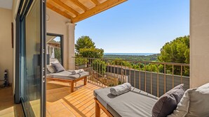 Ein Balkon mit Holzboden, zwei Betten mit Kissen, einer Glastür und Blick auf Bäume sowie eine entfernte Landschaft.