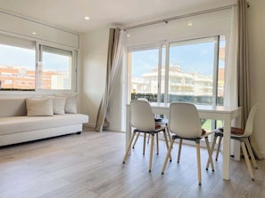 A modern living room with a sofa, dining table, chairs, and large windows with curtains.