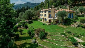 Une grande maison à deux étages, dotée d’un jardin bien entretenu et entourée d’arbres et de collines.