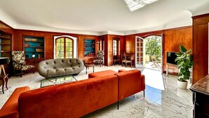 Un salon spacieux avec un grand canapé, des fauteuils et une table basse. On y trouve également des étagères à livres, un piano et un téléviseur. La pièce est dotée de grandes fenêtres et offre une vue sur l’extérieur.