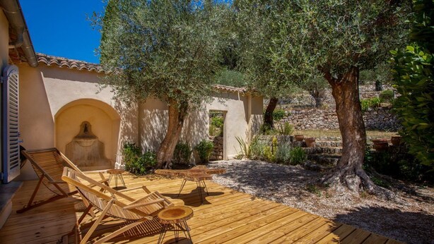 Une terrasse extérieure avec des meubles en bois, des oliviers et un mur en pierre.
