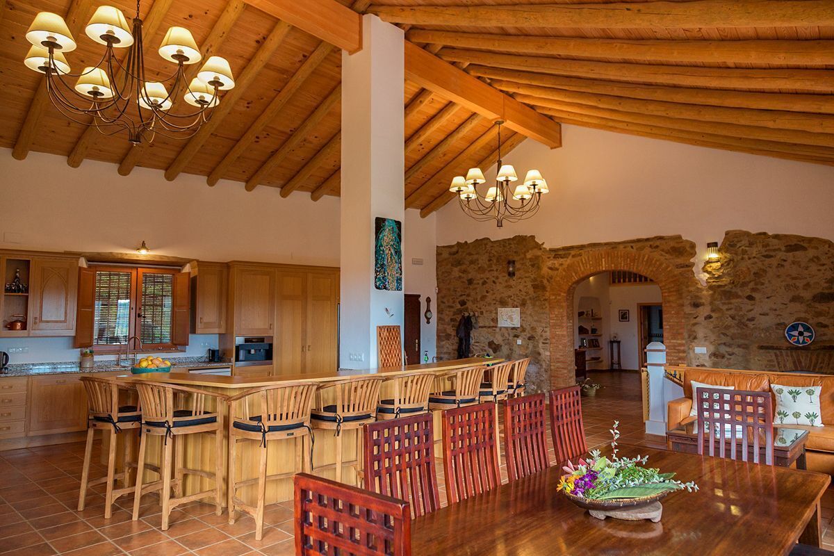 Una cocina amplia con armarios de madera y una isla central, un comedor con sillas de madera y una pared de piedra al fondo.