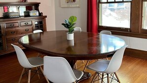 A dining area with a round wooden table, four white chairs, a potted plant, a chandelier, a cabinet with decorative items, and a window with a red curtain. - Historic Oak Park Residence (Oak Park)