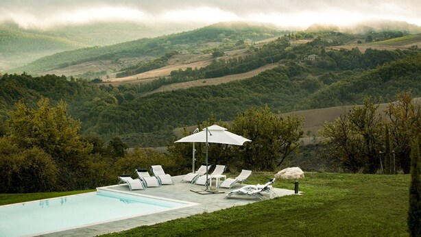 Zona de piscina con sillones de descanso y sombrillas, rodeada de un paisaje exuberante y colinas.