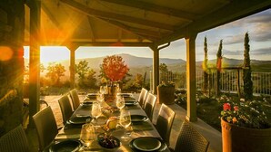Un área de comedor al aire libre con una mesa lista para una comida, rodeada de sillas, y vista a un paisaje pintoresco con montañas y árboles.