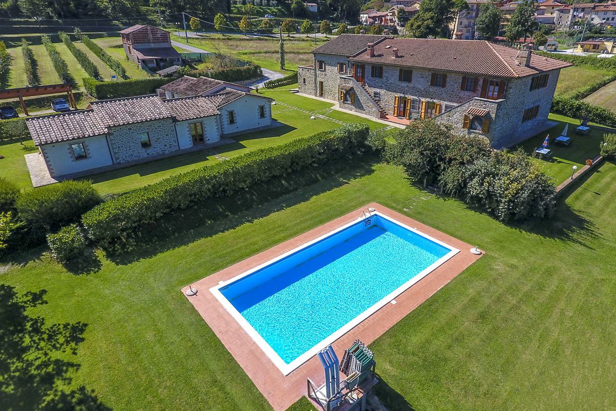 Una amplia casa de piedra con piscina, rodeada de césped cuidado y una cancha de tenis.