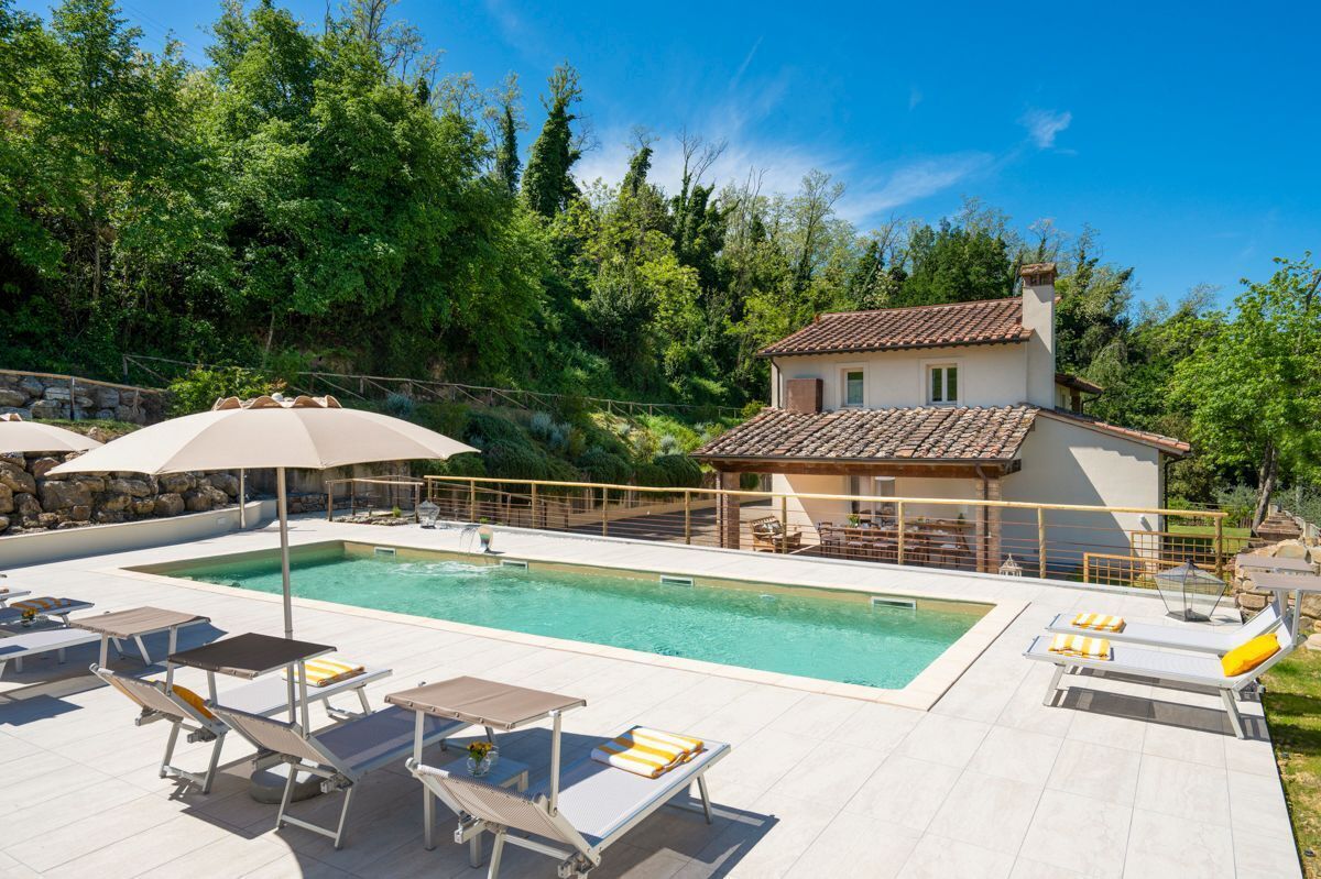 Un espace piscine agrémenté de chaises longues et de parasols, entouré d’un mur de pierre et d’arbres.