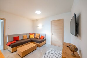 Un salon moderne avec un canapé d’angle, une table basse en bois, un tapis à motifs et un téléviseur à écran plat fixé au mur.