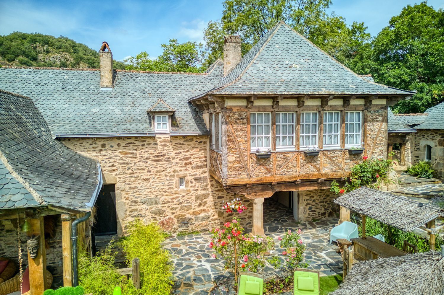 A stone building with a steeply pitched roof, a small balcony with windows, and a stone pathway leading to a seating area with chairs.