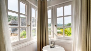 Una habitación con dos ventanas amplias, una mesa redonda blanca con cubierta de vidrio y vistas a un edificio tradicional y vegetación exterior.
