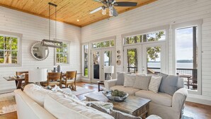 A spacious room with a high ceiling, wooden beams, a ceiling fan, and large windows offering a view of the outdoors.