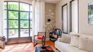 A living room with a white sofa, a wicker chair, a black coffee table with bowls, a stone wall, and a large window with curtains.