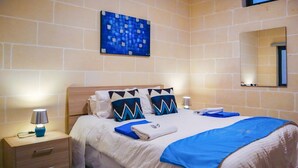 Een netjes opgemaakt bed met een blauwe deken en kussens, een bedlamp, een nachtkastje en een spiegel aan de muur.