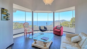 Un salón amplio con una mesa de centro de vidrio, un sofá rojo, una silla negra y una silla rosa. Amplias ventanas ofrecen vistas al mar y las colinas.