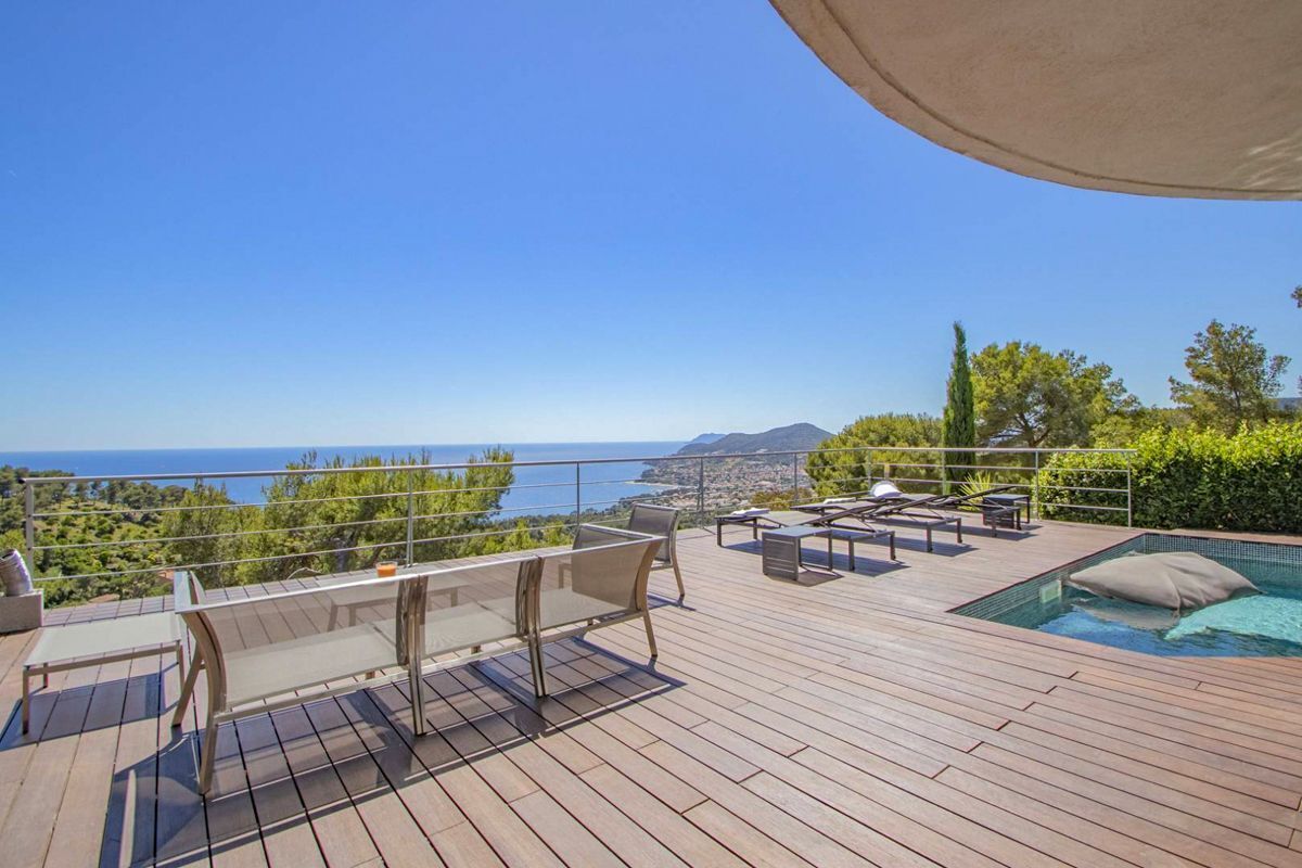 Une terrasse en bois avec mobilier d’extérieur, une piscine et une vue panoramique sur la mer et les montagnes.