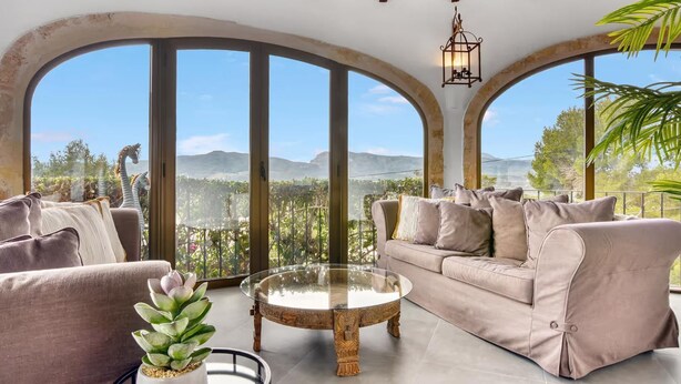 A spacious living room with a beige sofa, a glass coffee table, and large arched windows offering a view of a mountainous landscape.