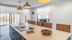 A modern kitchen with a central island, wicker chairs, pendant lighting, and large windows.