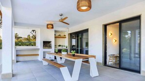 Une terrasse extérieure avec une table et des bancs en bois, un ventilateur de plafond et une porte vitrée donnant sur un balcon.