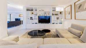 Un salon moderne avec un grand téléviseur à écran plat, un canapé beige et une table basse. On y trouve des étagères ornées d’objets décoratifs et une bibliothèque.