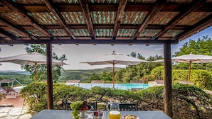 Ein überdachter Außenbereich mit einem Tisch für zwei Personen, Blick auf einen Swimmingpool und üppiges Grün im Hintergrund.