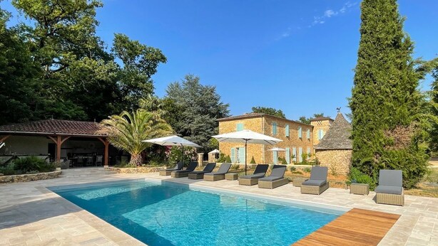 A swimming pool with lounge chairs and umbrellas, surrounded by stone buildings and greenery. - Domaine Des Maronniers (Carsac-Aillac)