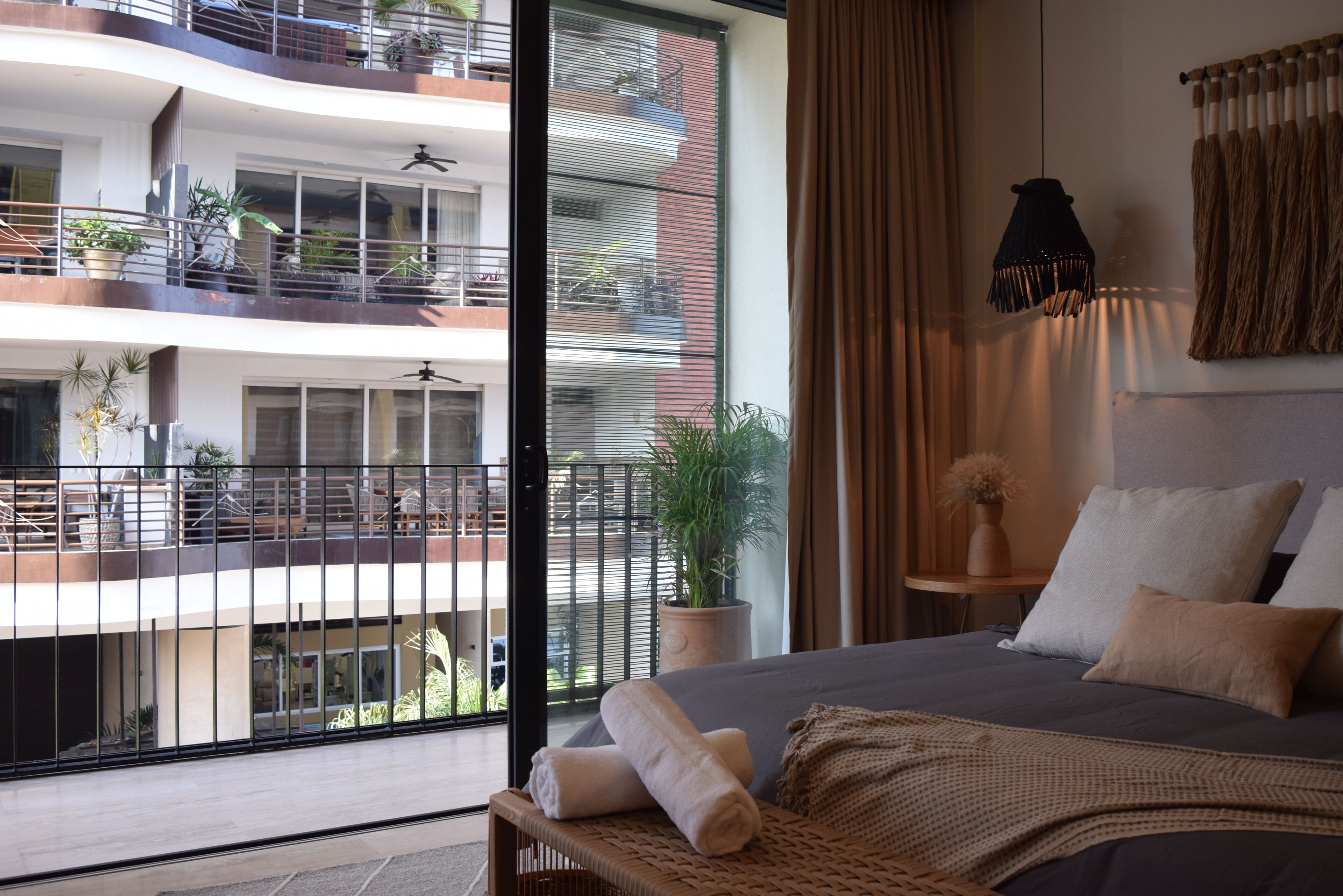 Une chambre d’hôtel avec un grand lit, une plante en pot, un luminaire suspendu et une vue sur les balcons extérieurs de l’immeuble.