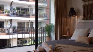 A hotel room with a large bed, a potted plant, a hanging light fixture, and a view of the building's exterior balconies.