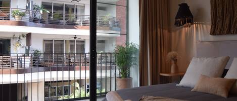 A hotel room with a large bed, a potted plant, a hanging light fixture, and a view of the building's exterior balconies.