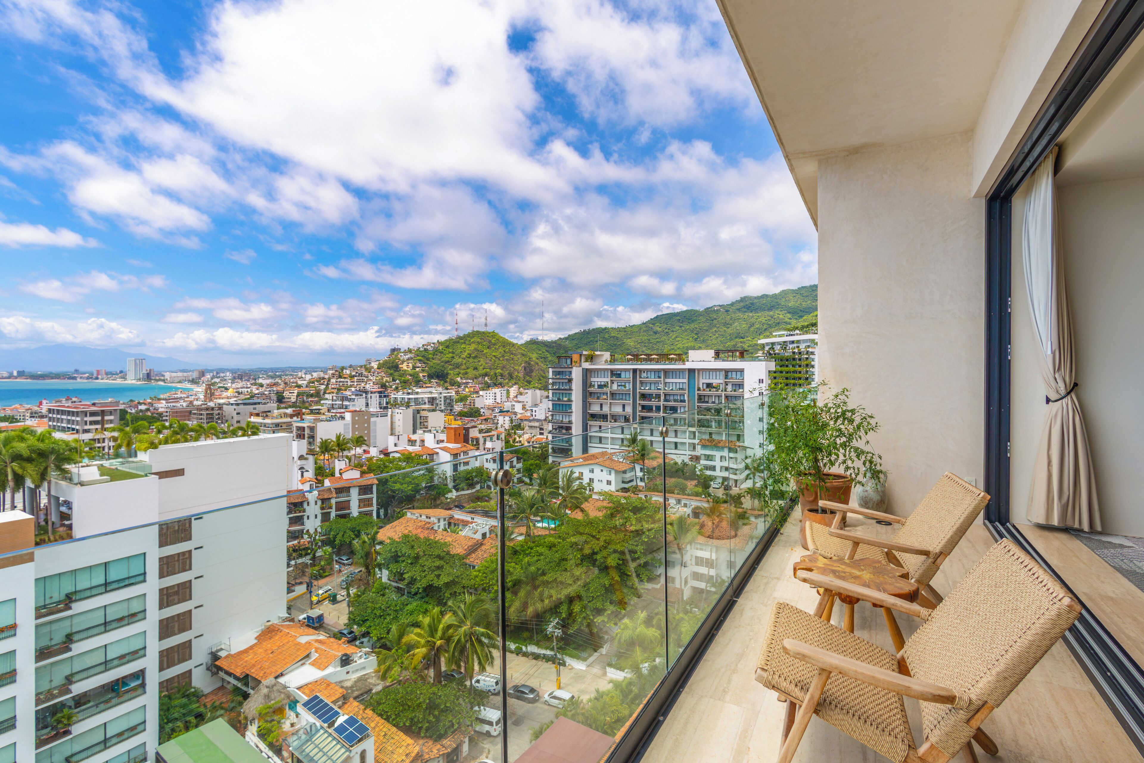 Ein Balkon mit Korbstühlen, einer Topfpflanze und Blick auf eine Stadtlandschaft mit Gebäuden und einem Strand.