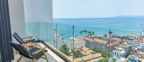 Ein Balkon mit zwei Holzstühlen, einer Glasreling und Blick auf das Meer und die Stadtlandschaft.