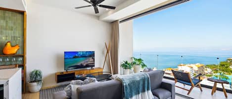 A modern living room with a large TV, a sofa, a ceiling fan, and a view of the ocean.