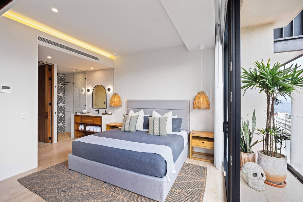 A modern bedroom with a large bed, bedside lamps, a vanity, and a potted plant.