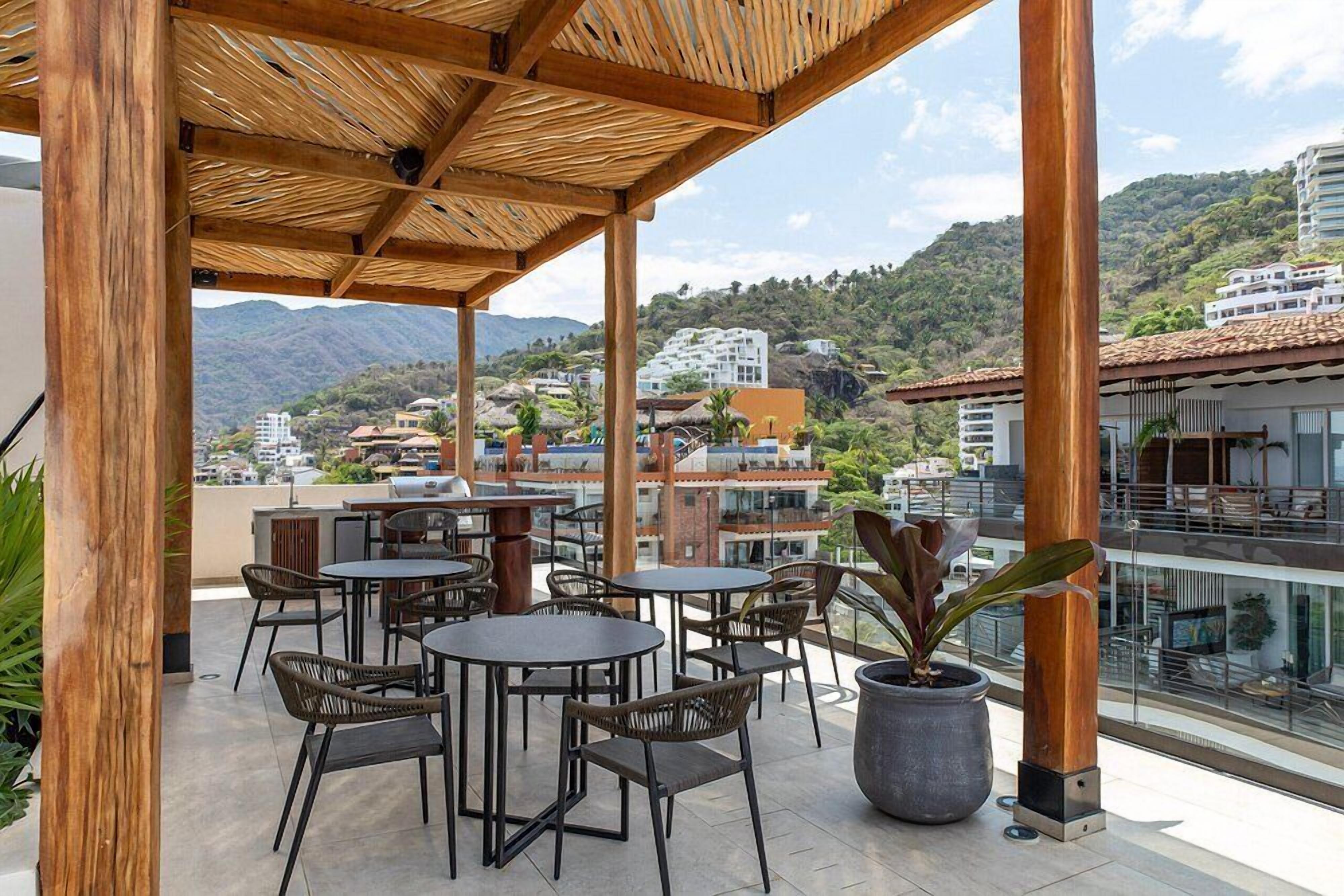 An outdoor patio with tables and chairs, a large potted plant, and a view of a hillside with buildings.