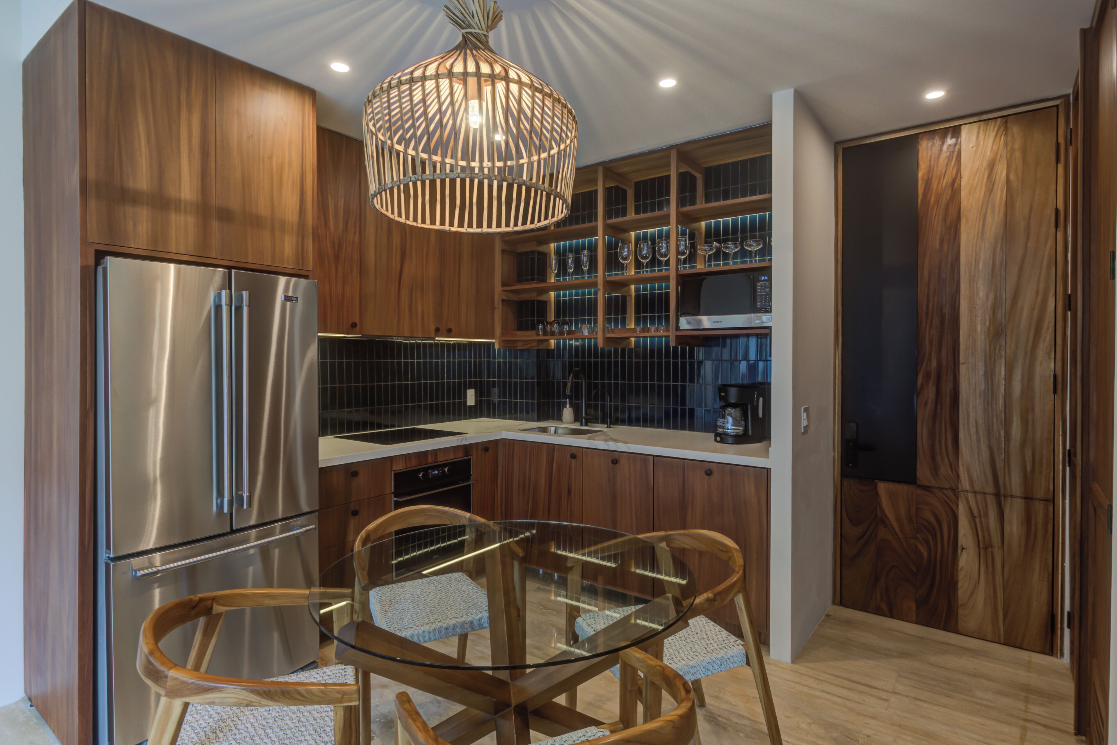 A modern kitchen with wooden cabinets, a stainless steel refrigerator, a glass dining table, and a hanging light fixture.