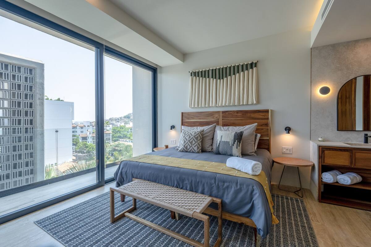A modern bedroom with a large window, a wooden headboard, and a bedside table with towels.